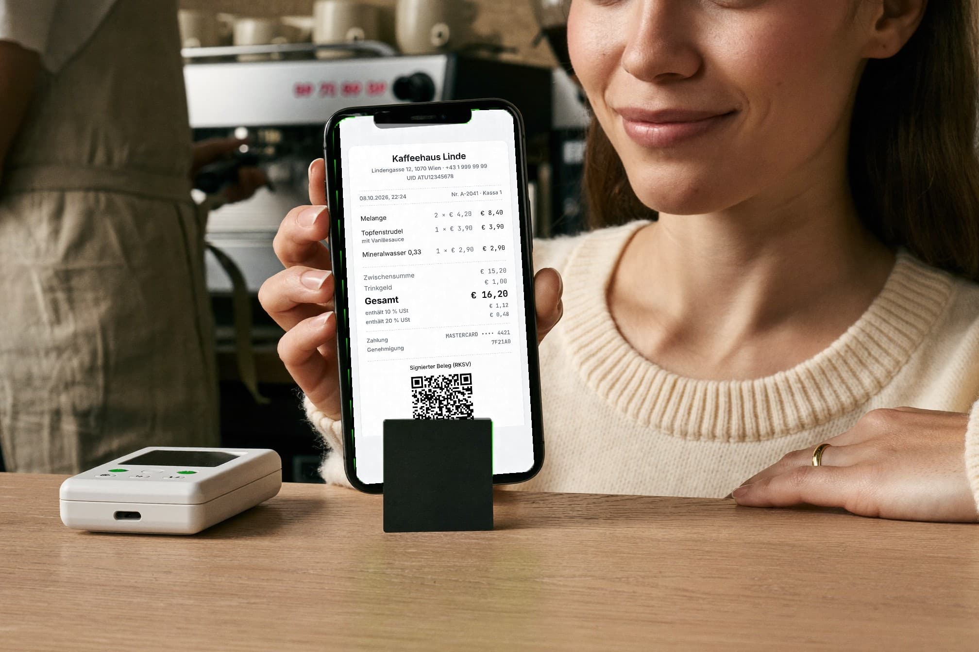 Eine Kundin an der Theke eines Kaffeehauses zeigt ihr iPhone mit dem digitalen Beleg: Kaffeehaus Linde, Melange, Topfenstrudel, Mineralwasser, Gesamt 16,20 Euro, RKSV-QR-Code. Vor ihr die kleine schwarze Kachel neben dem Kartenterminal.