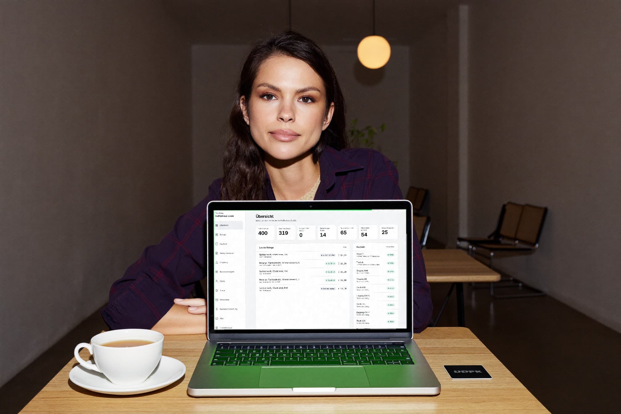 Eine Café-Besitzerin sitzt nach Ladenschluss am Tisch, der Laptop zeigt die Übersicht im Konto mit den Zahlen des Tages, daneben Tee und eine Kachel.